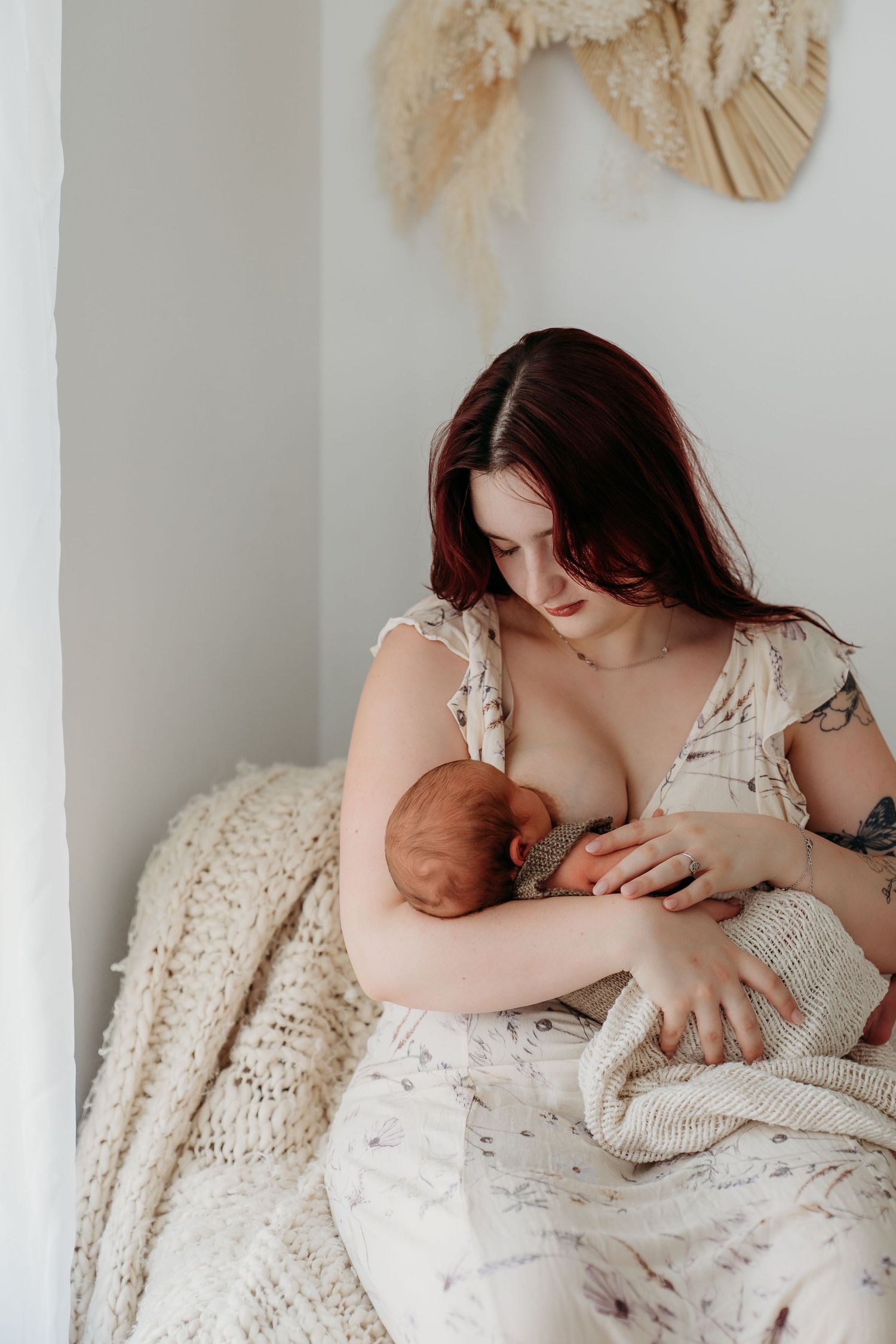 A mother with red hair breastfeeding her newborn baby while sitting on a bed with a cozy knit blanket.