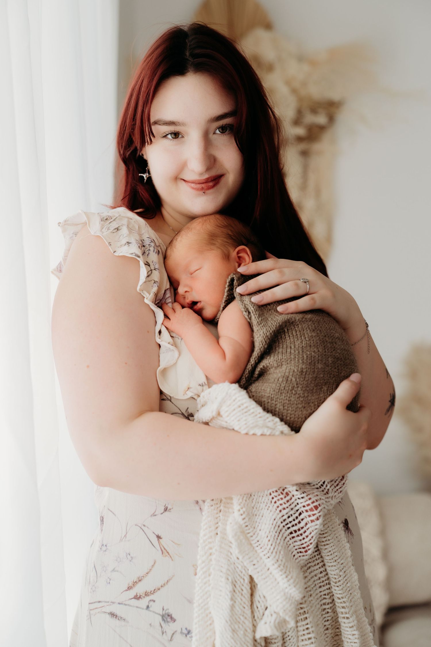 Young mother with red hair smiling while gently holding her sleeping newborn baby wrapped in a knit blanket.