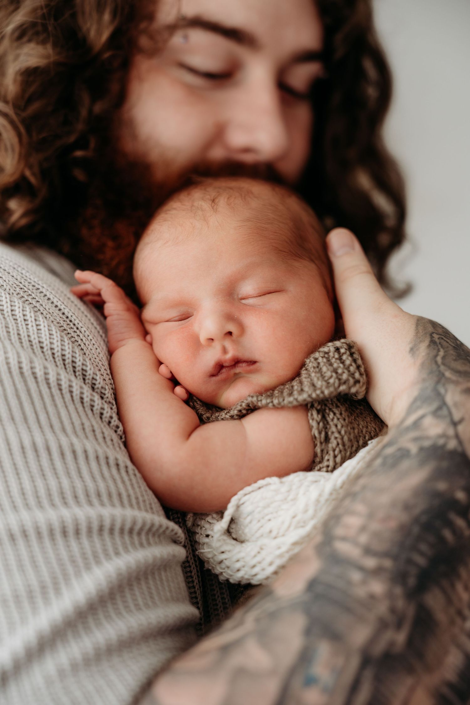 Tattooed father with curly hair gently holds sleeping newborn baby wrapped in knit blanket close to his chest.