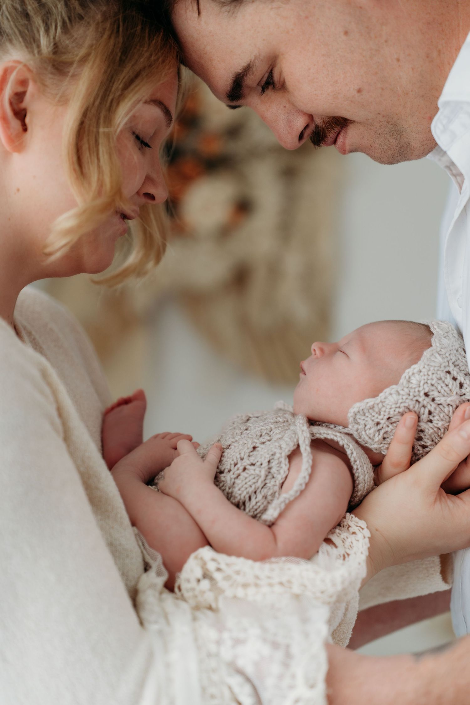 New parents tenderly hold their sleeping newborn wrapped in a knit outfit, foreheads touching in an intimate moment.