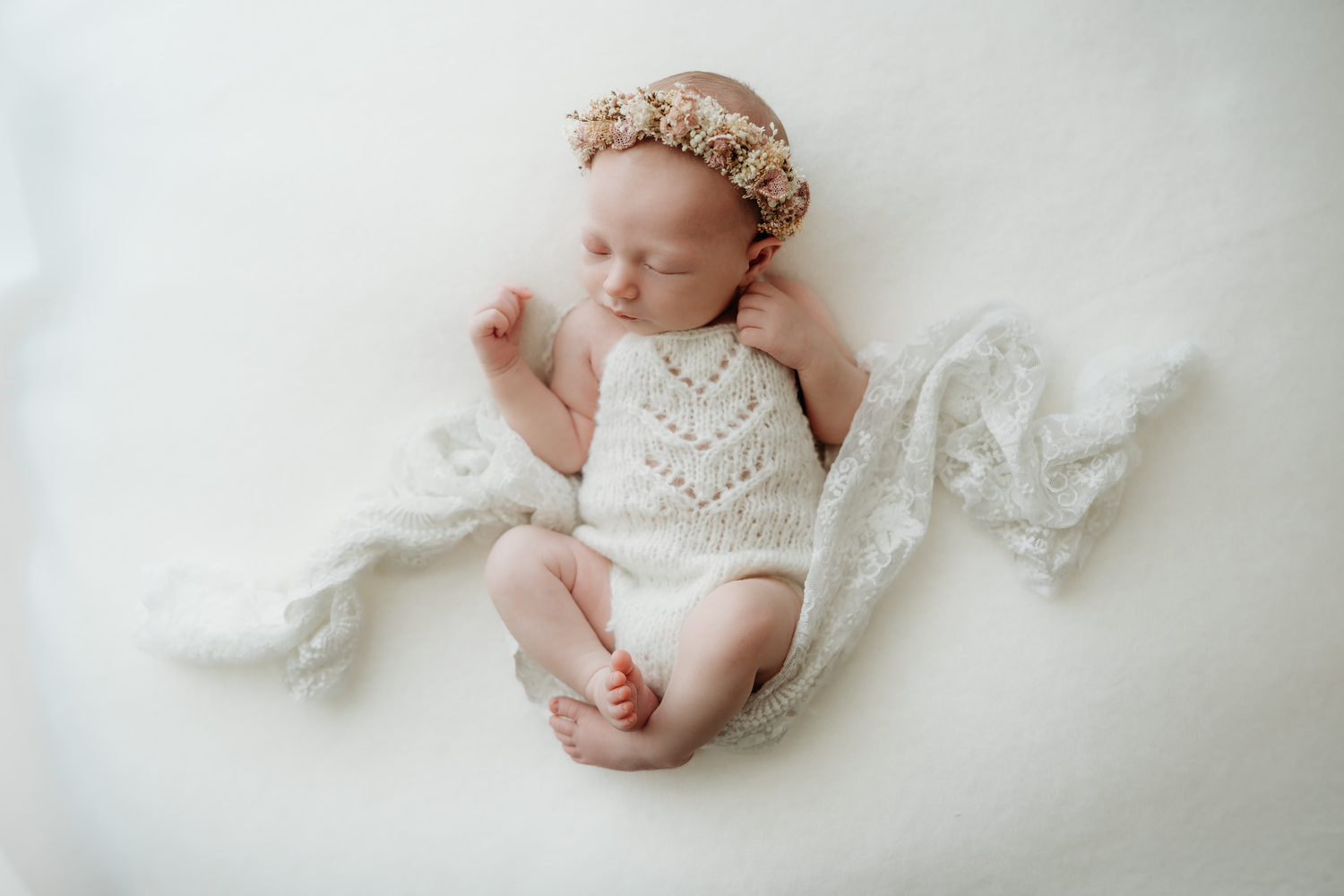 Sleeping newborn baby girl in white knit romper and floral headband on soft white blanket.