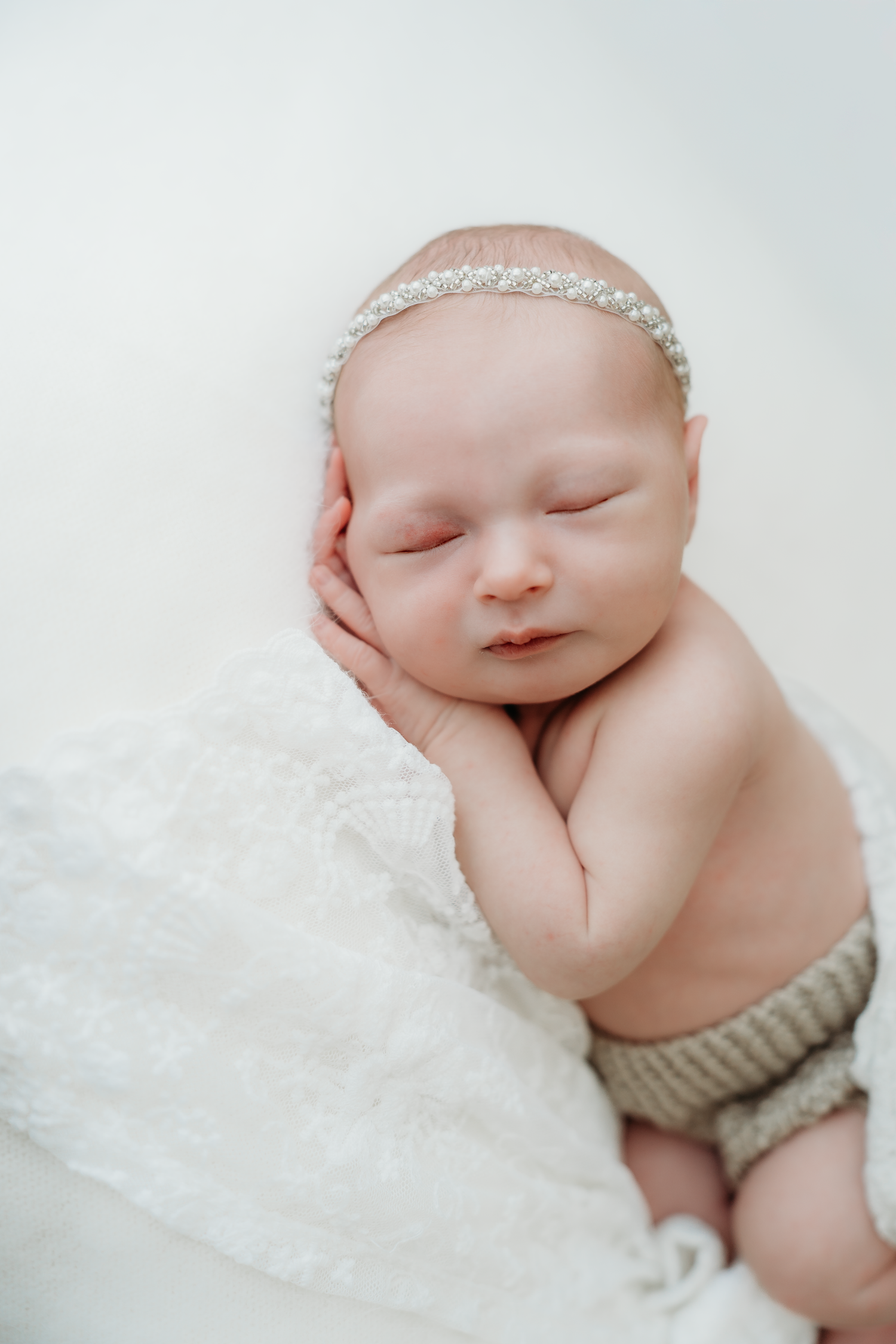 Sleeping newborn baby wearing a pearl headband and knit shorts, resting on a white lace blanket.