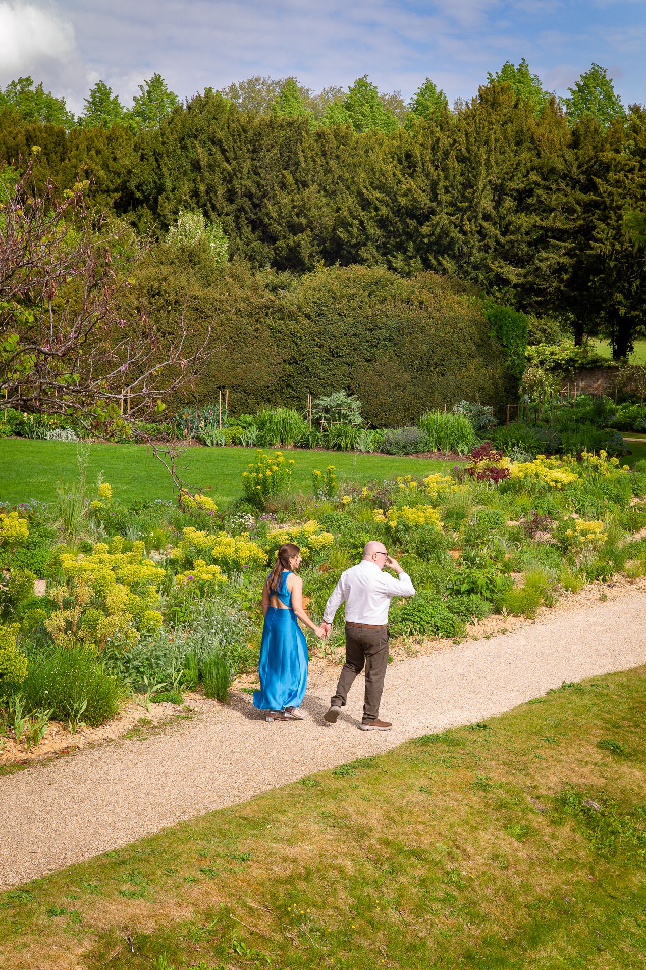 Couple holding hands walking along a garden path surrounded by lush greenery and yellow flowers on a sunny day.