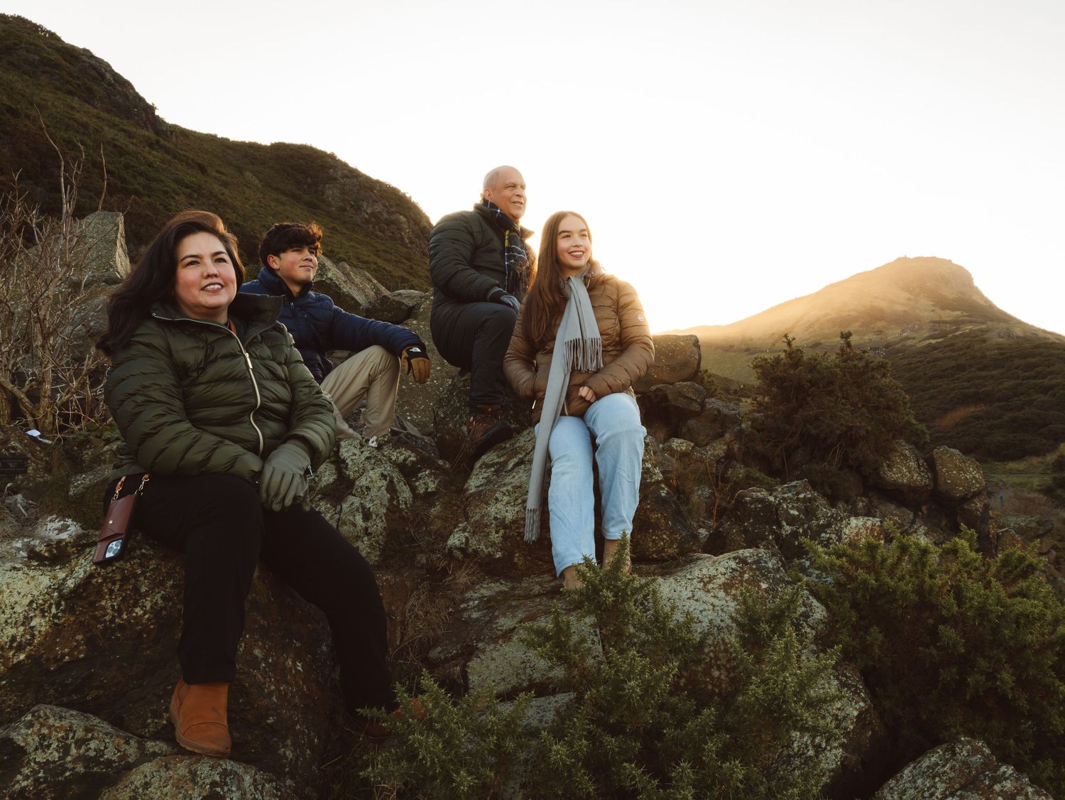 Four friends sit on rocky mountain terrain at golden hour, wearing warm winter jackets and scarves during a scenic hike.