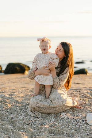 A young mother sits on a sandy beach holding her smiling baby girl in a floral dress at golden hour.