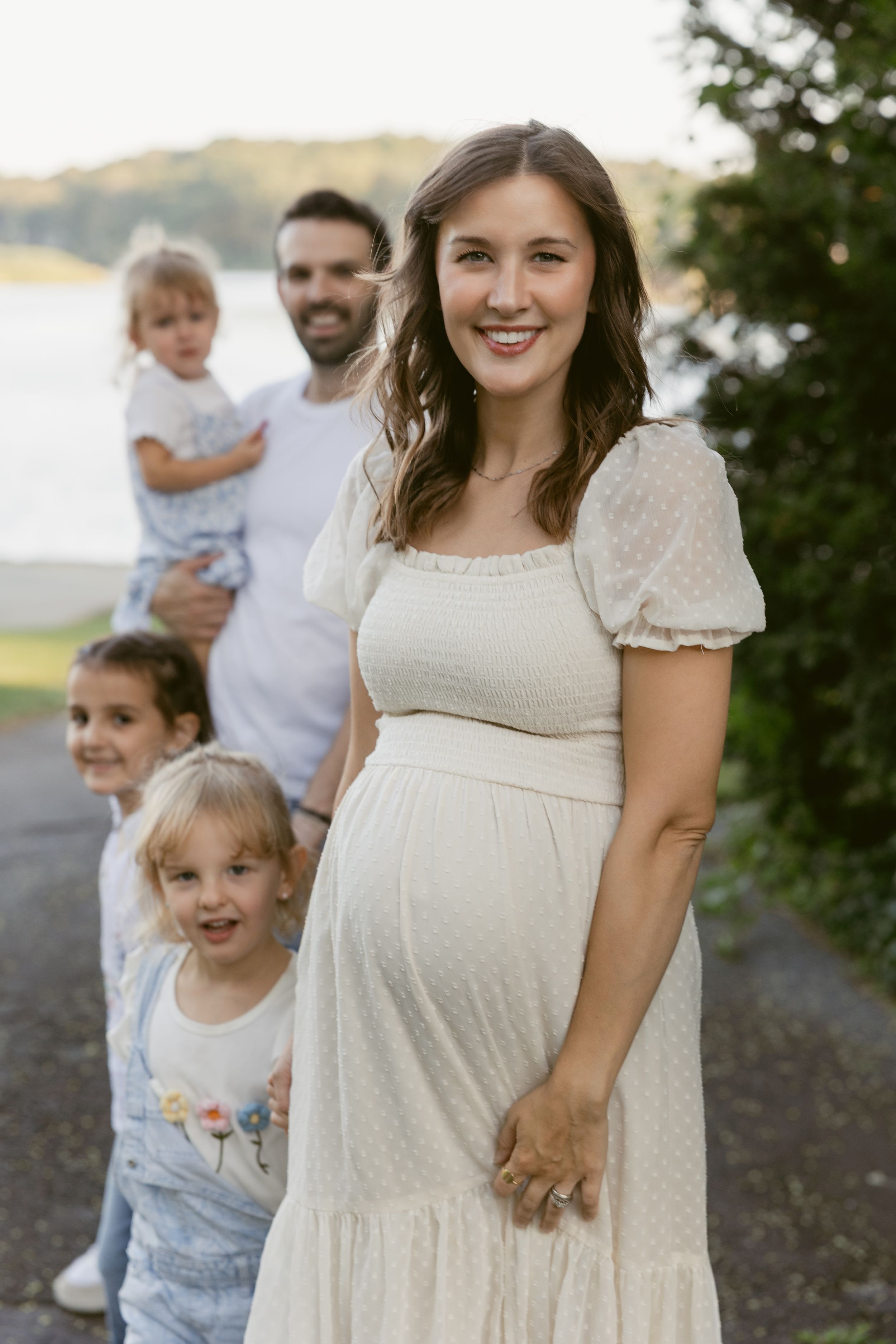 Pregnant woman in white dress smiles outdoors with husband holding toddler and two young children beside a lake.