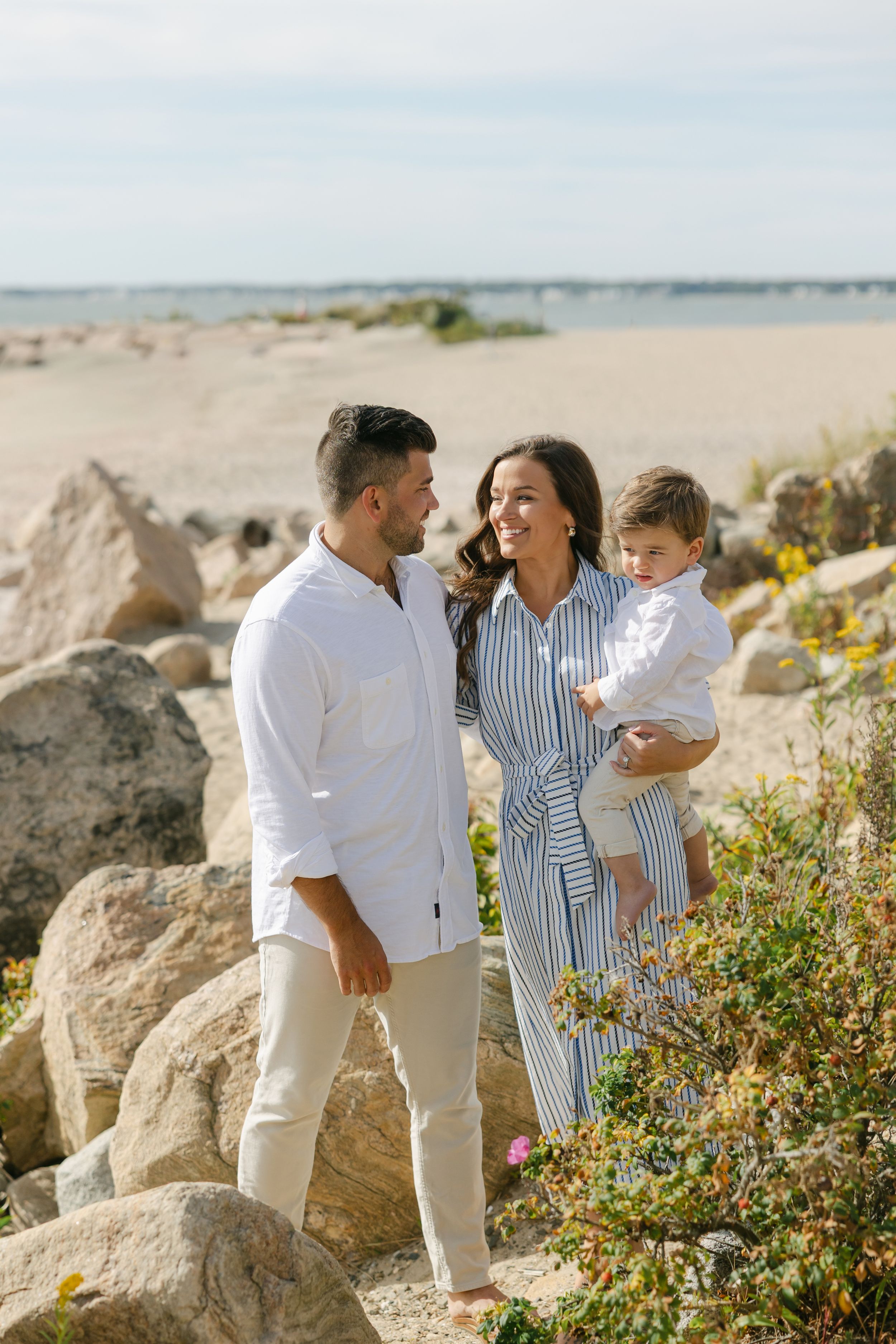 A young family of three poses on rocky beach, parents smiling at each other while mother holds toddler.