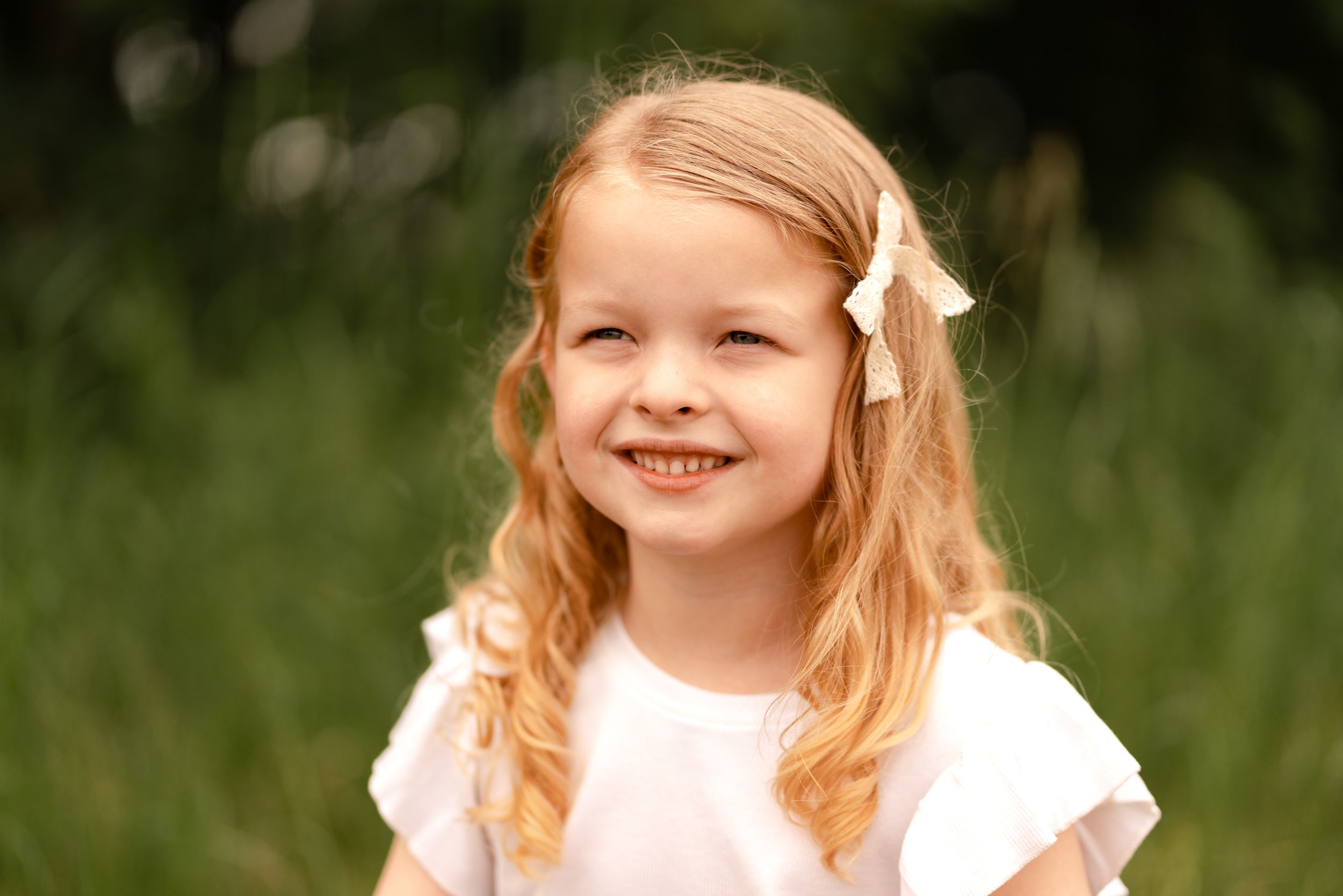 Smiling young girl with blonde hair and white bow outdoors in green nature setting, wearing white dress.