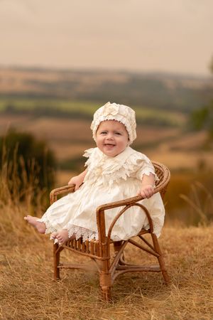 Smiling baby in white lace dress and bonnet sitting in wicker chair outdoors in golden field.