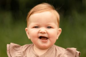Smiling baby girl with red hair wearing a pink ruffle dress, laughing outdoors with green bokeh background.