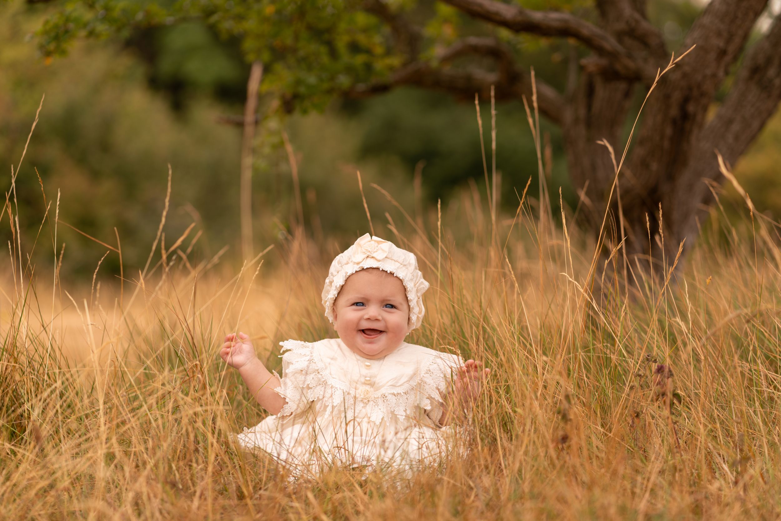 Smiling baby in white dress and bonnet sitting in golden tall grass field with trees in background.