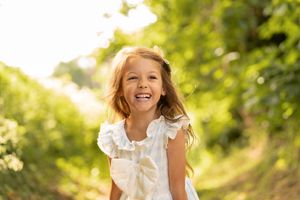 A joyful young girl in a white dress laughs outdoors on a sunny green path, bathed in warm golden sunlight.