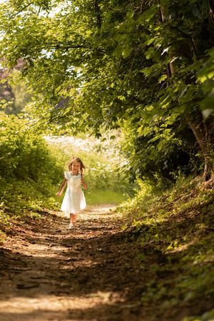Young girl in white dress running joyfully down a sunlit forest path surrounded by lush green trees.