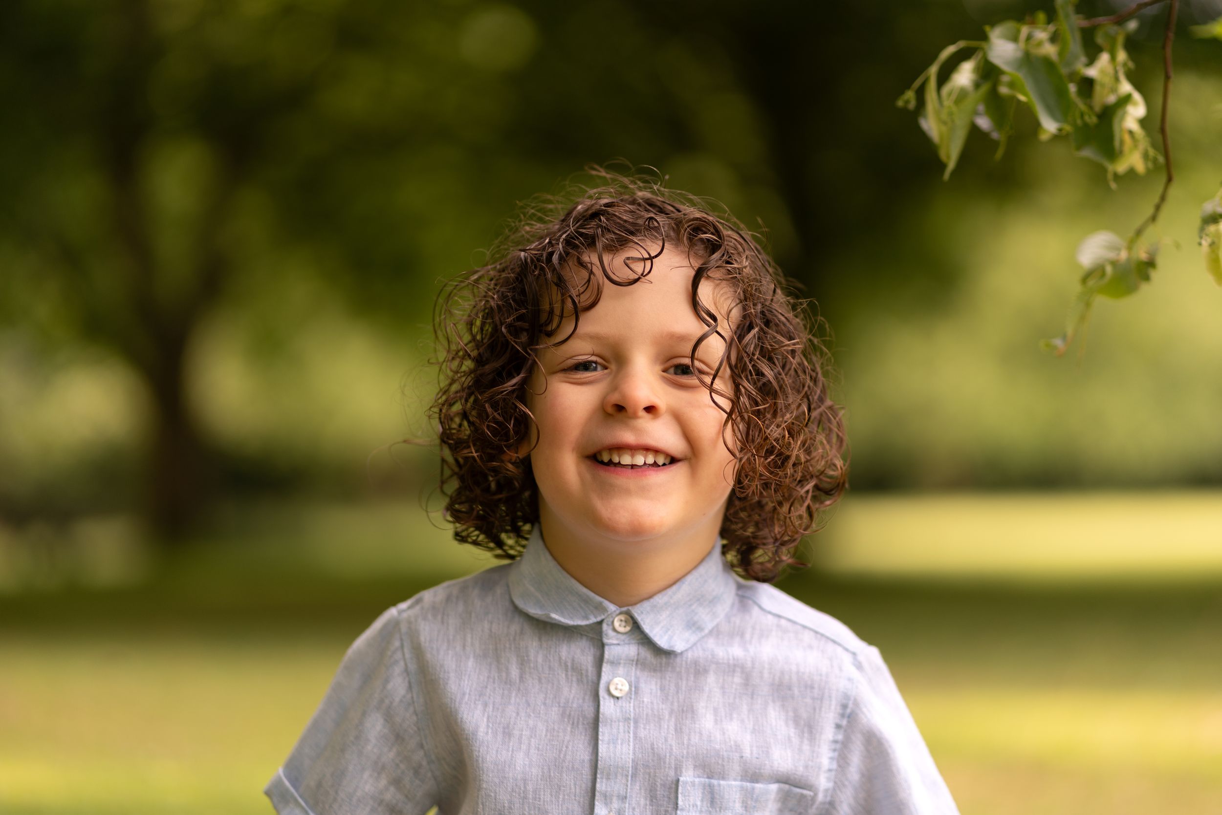 Smiling curly-haired boy in a light blue linen shirt posing outdoors in a lush green park.