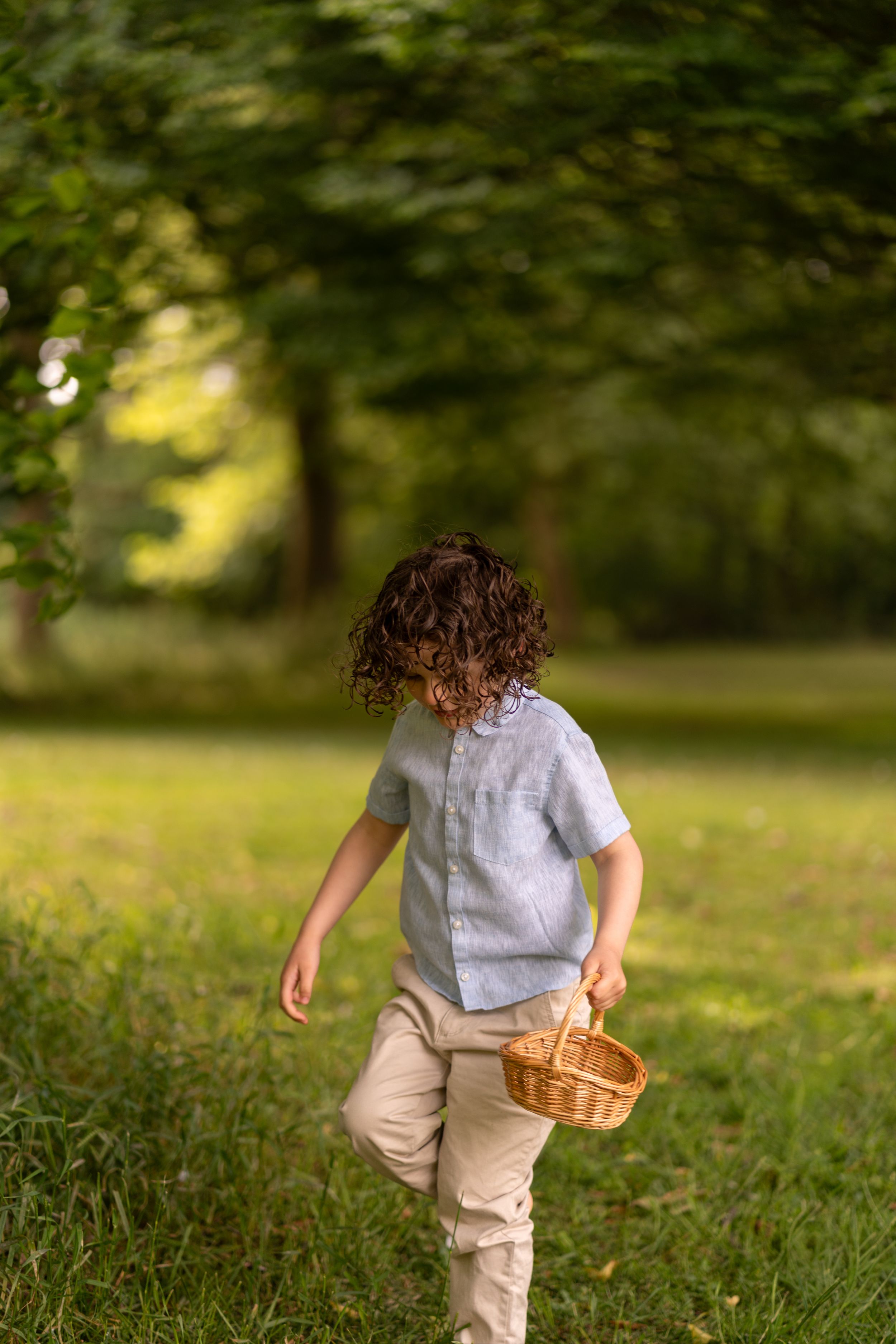 Curly-haired child running through a green park carrying a wicker basket on a sunny day.