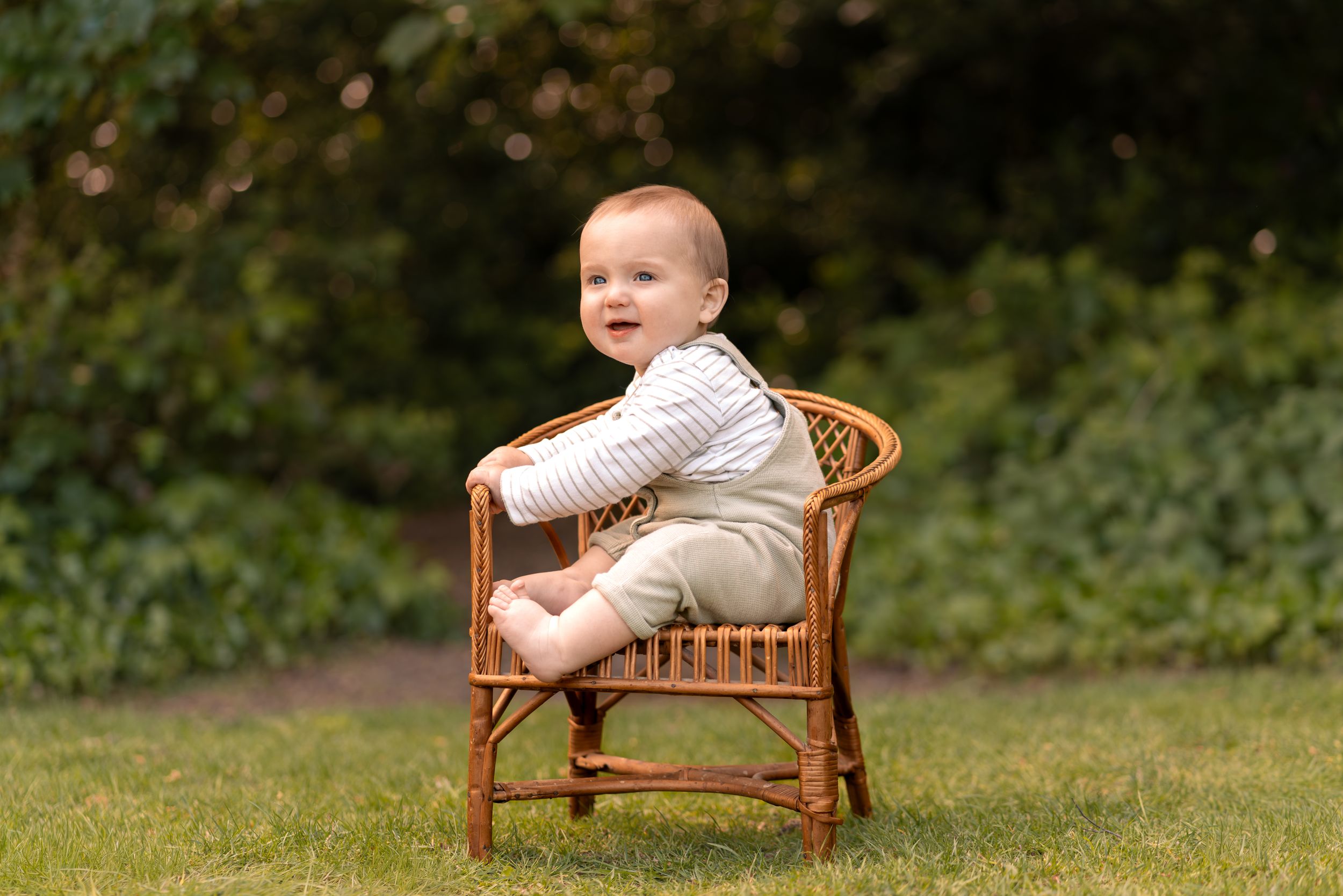 Adorable baby sitting in a small wicker chair on green grass outdoors during a childhood photography session in Lincolnshire.