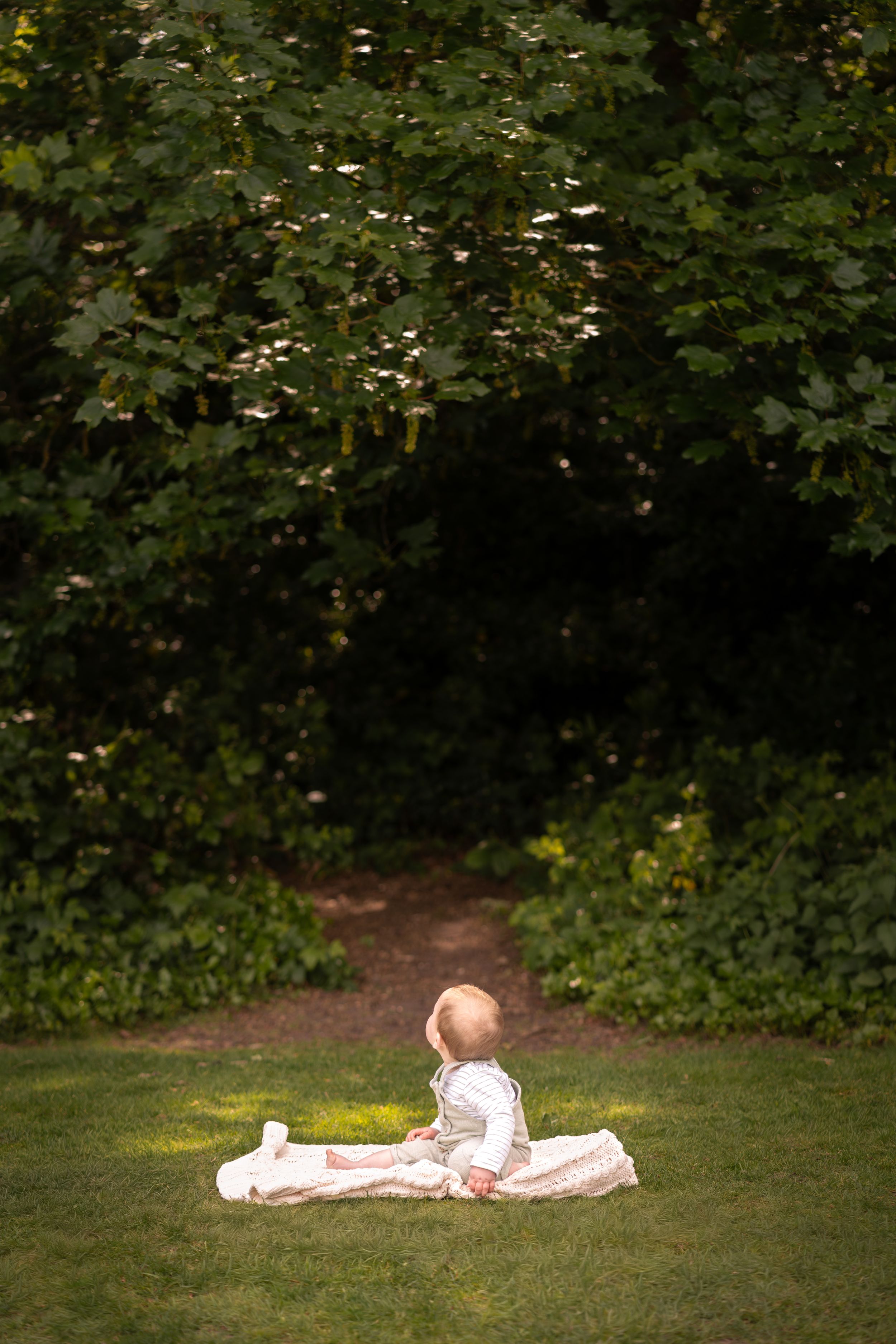 Baby in white outfit sitting on a blanket on green grass surrounded by lush trees in a serene garden setting.
