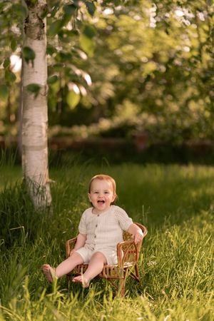 Happy baby in white romper sitting in wicker chair on green grass under birch tree in sunny garden.