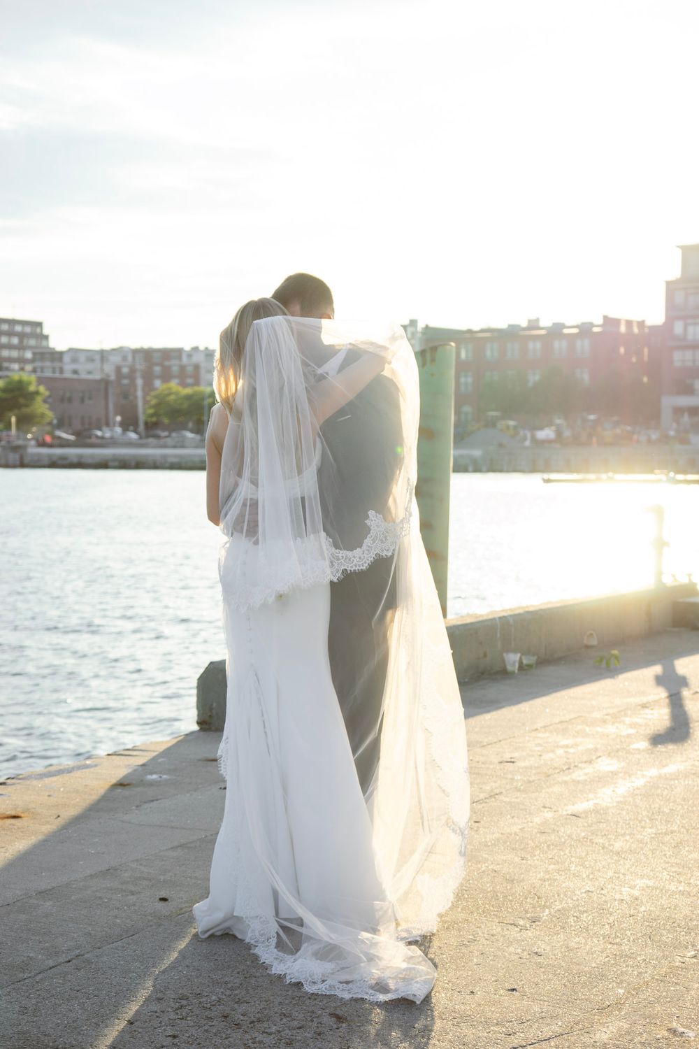 Bride and groom share a tender kiss by the waterfront at sunset, her lace-trimmed veil cascading down.