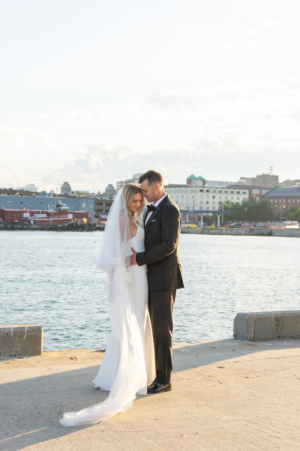Bride and groom embracing on a waterfront pier with a scenic harbor and city skyline in the background.