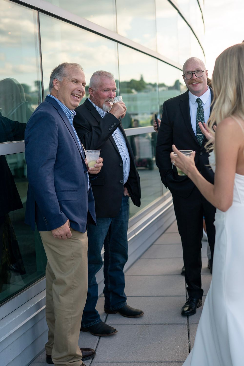 Group of men in formal attire laughing and drinking at outdoor reception.