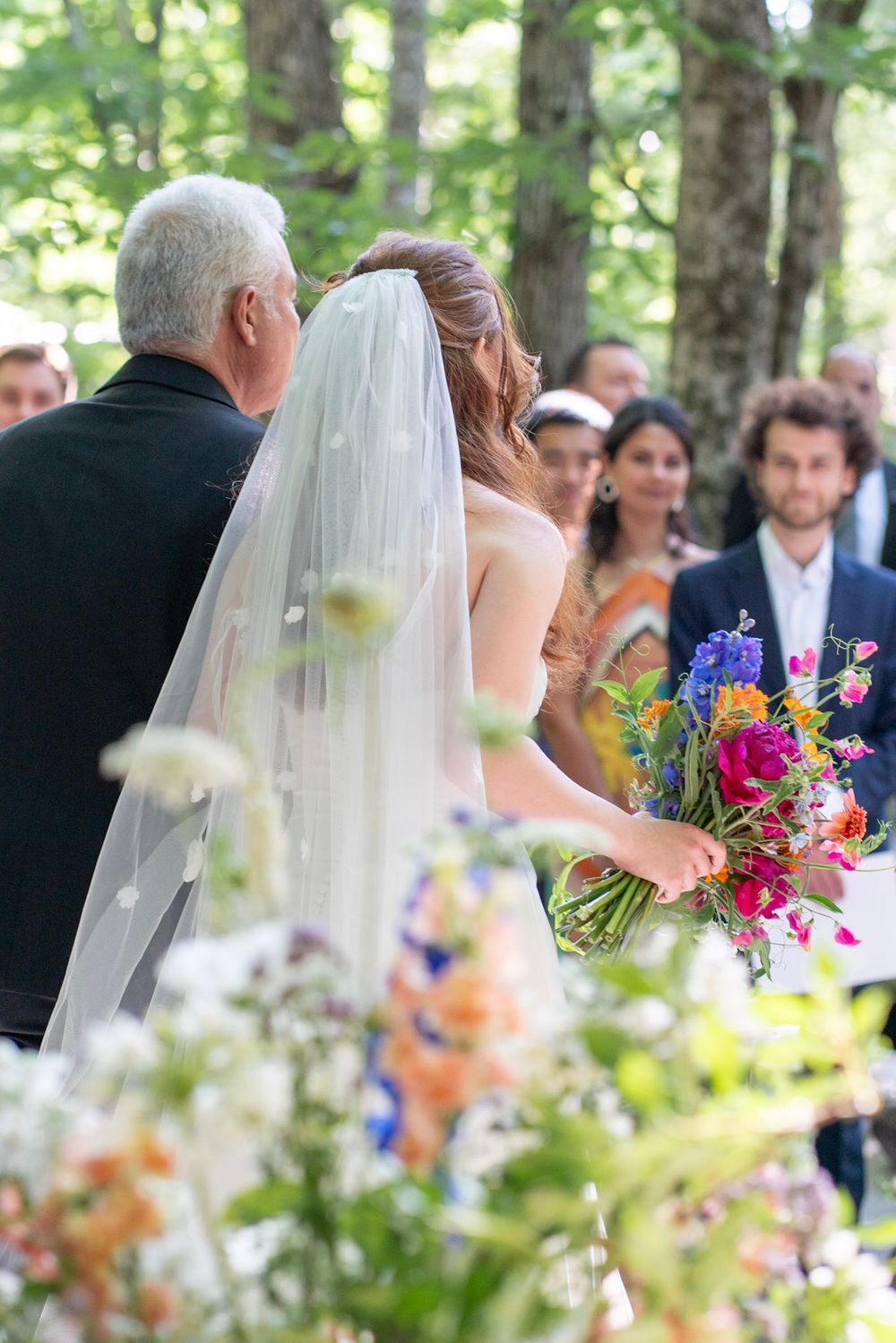 Bride in white gown and veil holds colorful bouquet while walking down outdoor aisle with older gentleman.