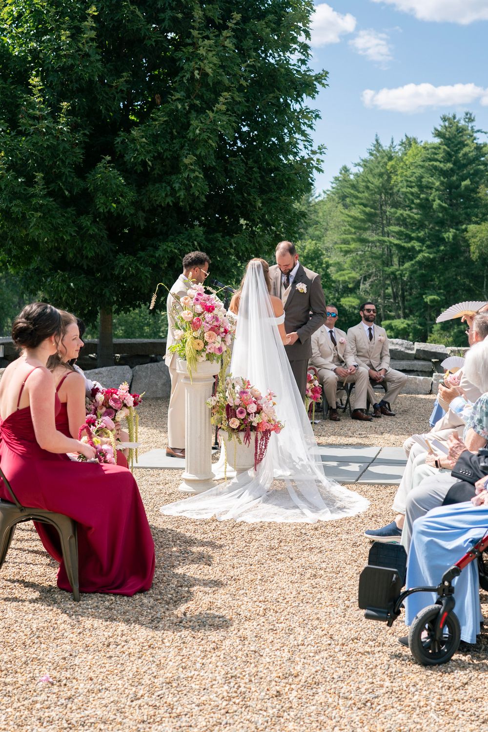 Outdoor wedding ceremony with bride and groom at altar, bridesmaid in red gown seated, guests watching on sunny day.