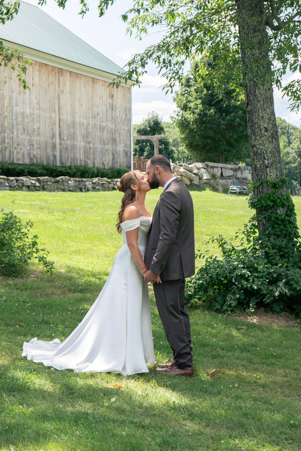 Bride and groom sharing romantic kiss holding hands on green lawn beside rustic barn and stone wall.