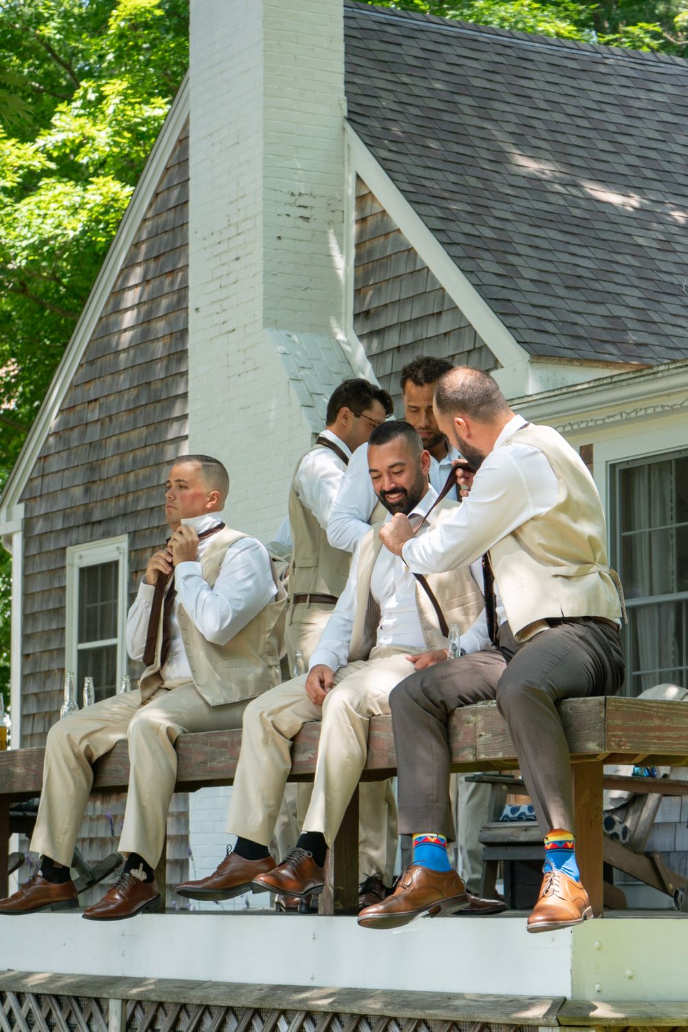 Groomsmen helping each other with ties and suspenders while sitting on porch steps before wedding ceremony.