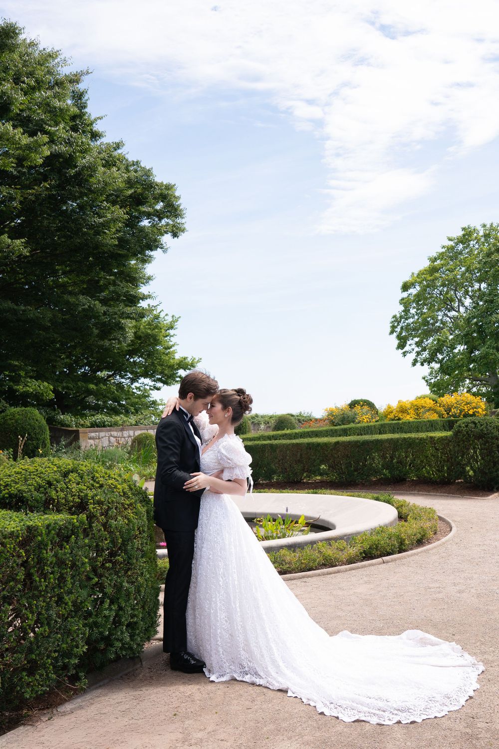 Bride and groom share a romantic kiss in a formal garden, bride wearing elegant white lace gown with long train.