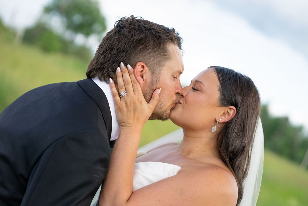 A bride and groom share a romantic kiss outdoors on their wedding day, with green fields in the background.