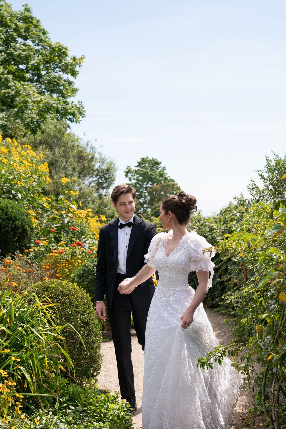 A bride and groom walk hand in hand through a lush garden path, smiling at each other on their wedding day.