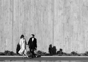 Elegant couple in formal attire holding hands while walking along a wooden wall in a black and white photo.