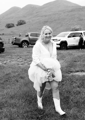 Bride in white dress and boots holding bouquet, laughing outdoors with hills and trucks in background.