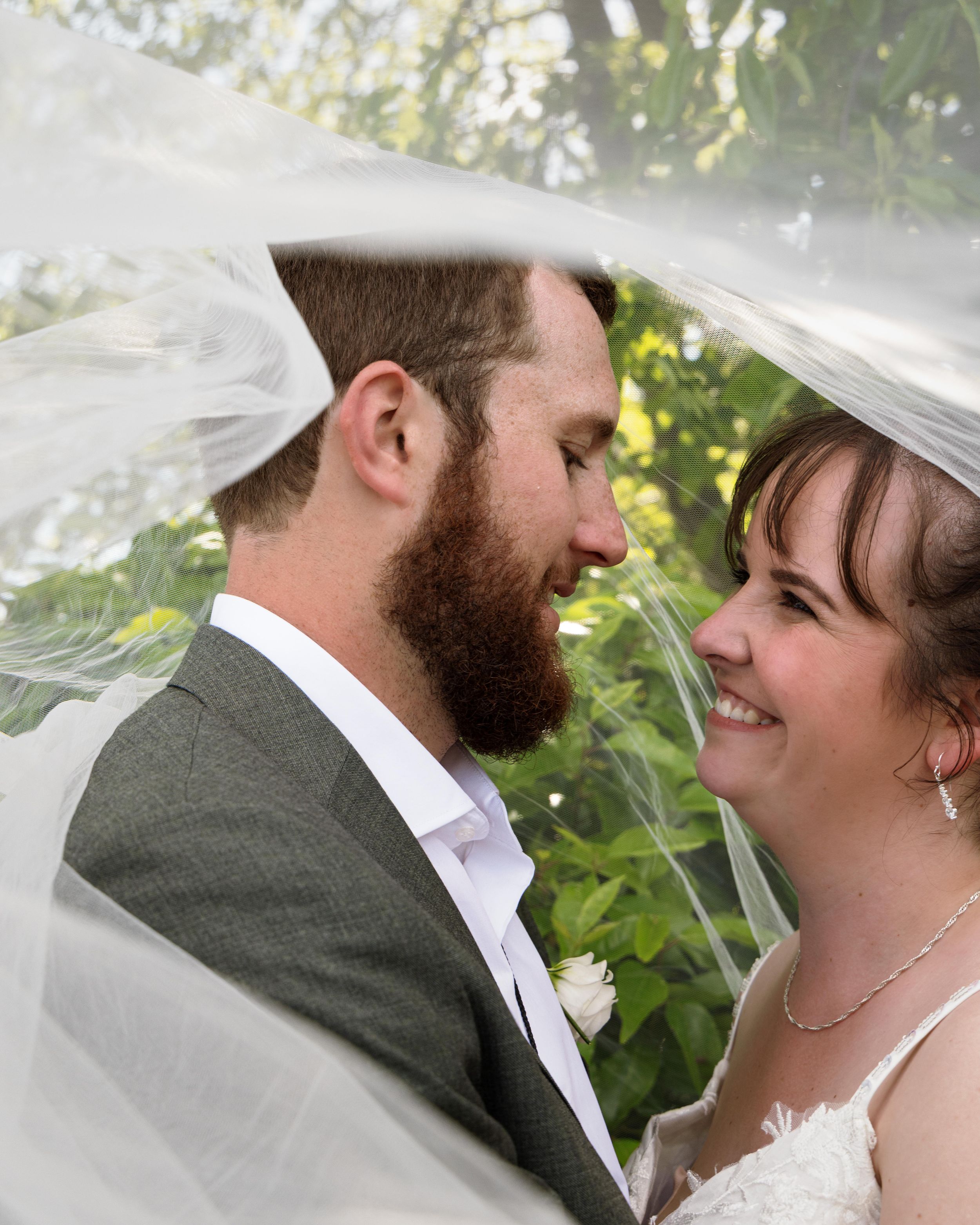 Bride and groom share a romantic moment under a flowing white veil outdoors on their wedding day.
