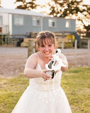 Smiling bride in white lace gown holding a toy horse figurine outdoors at a farm venue during golden hour.