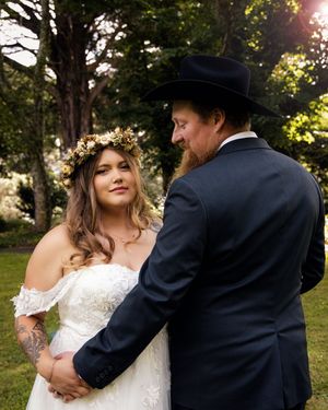 Bride in floral crown and white off-shoulder gown stands with groom in black cowboy hat and suit outdoors.