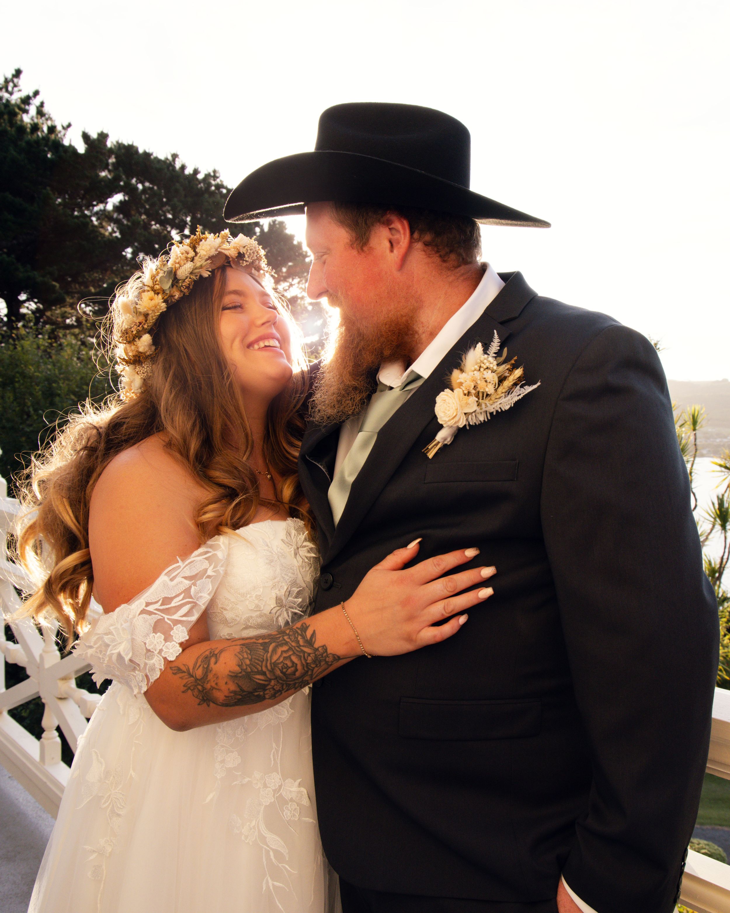 Bride in floral crown and groom in black cowboy hat share a romantic moment in wedding attire outdoors.