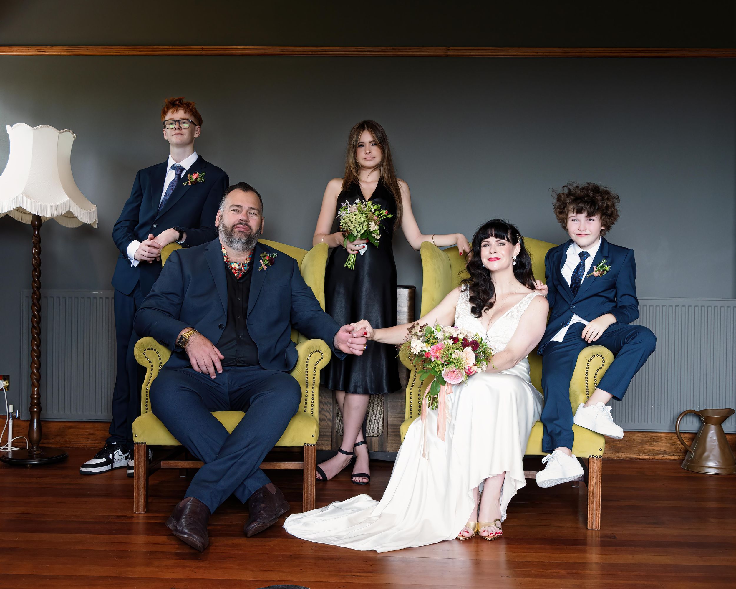 Wedding party of five posing formally, bride in white gown holding bouquet, groom and guests in navy suits on yellow chairs.