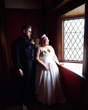 Bride and groom stand by a diamond-paned window in a dark red room, she wears a white dress and pink hair accessory.