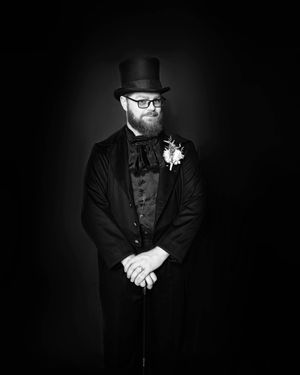Black and white portrait of bearded man in top hat, bow tie, and formal coat with boutonniere, holding a cane.
