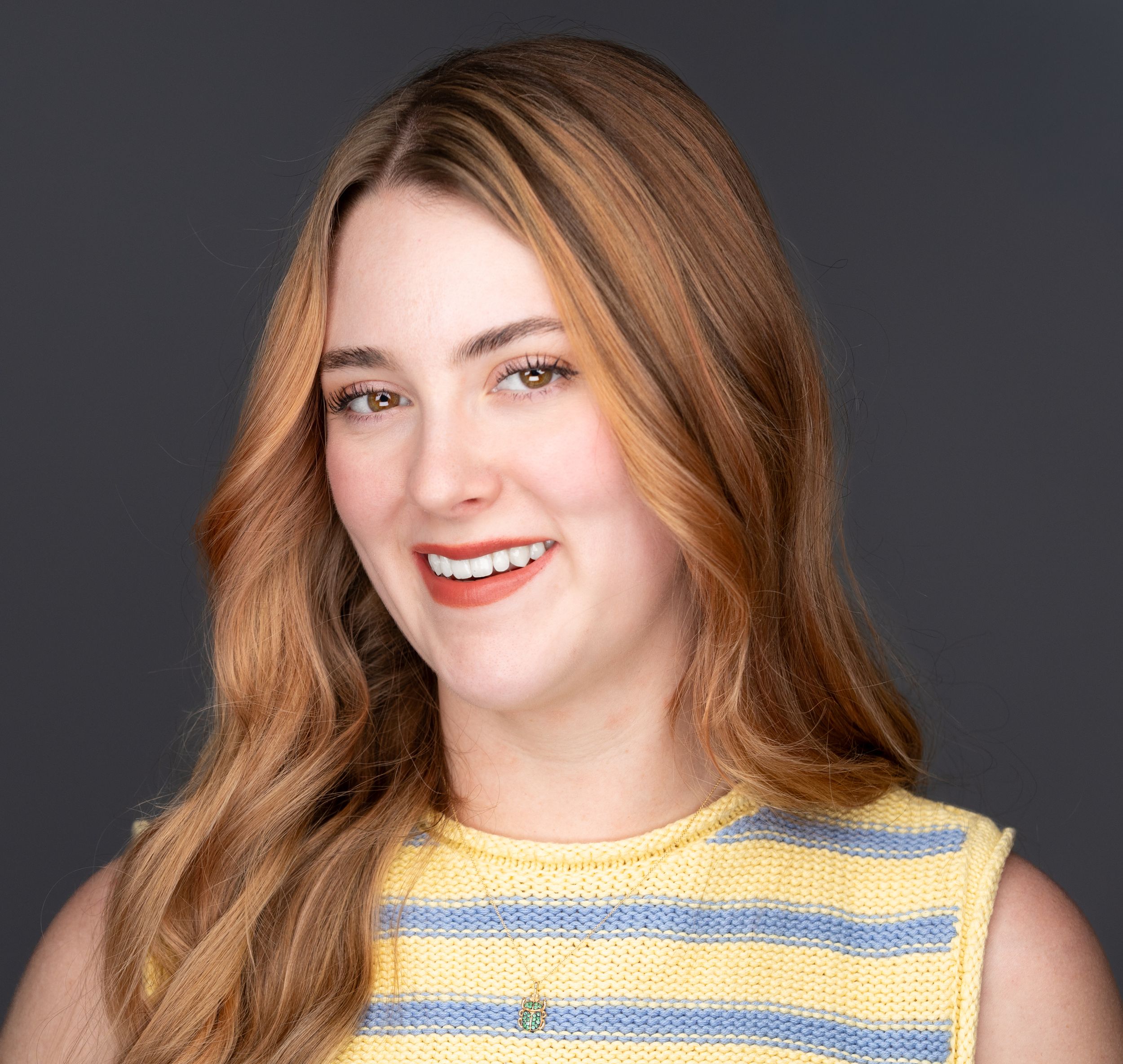 Young woman with long auburn hair smiling, wearing yellow striped sleeveless top against dark background.