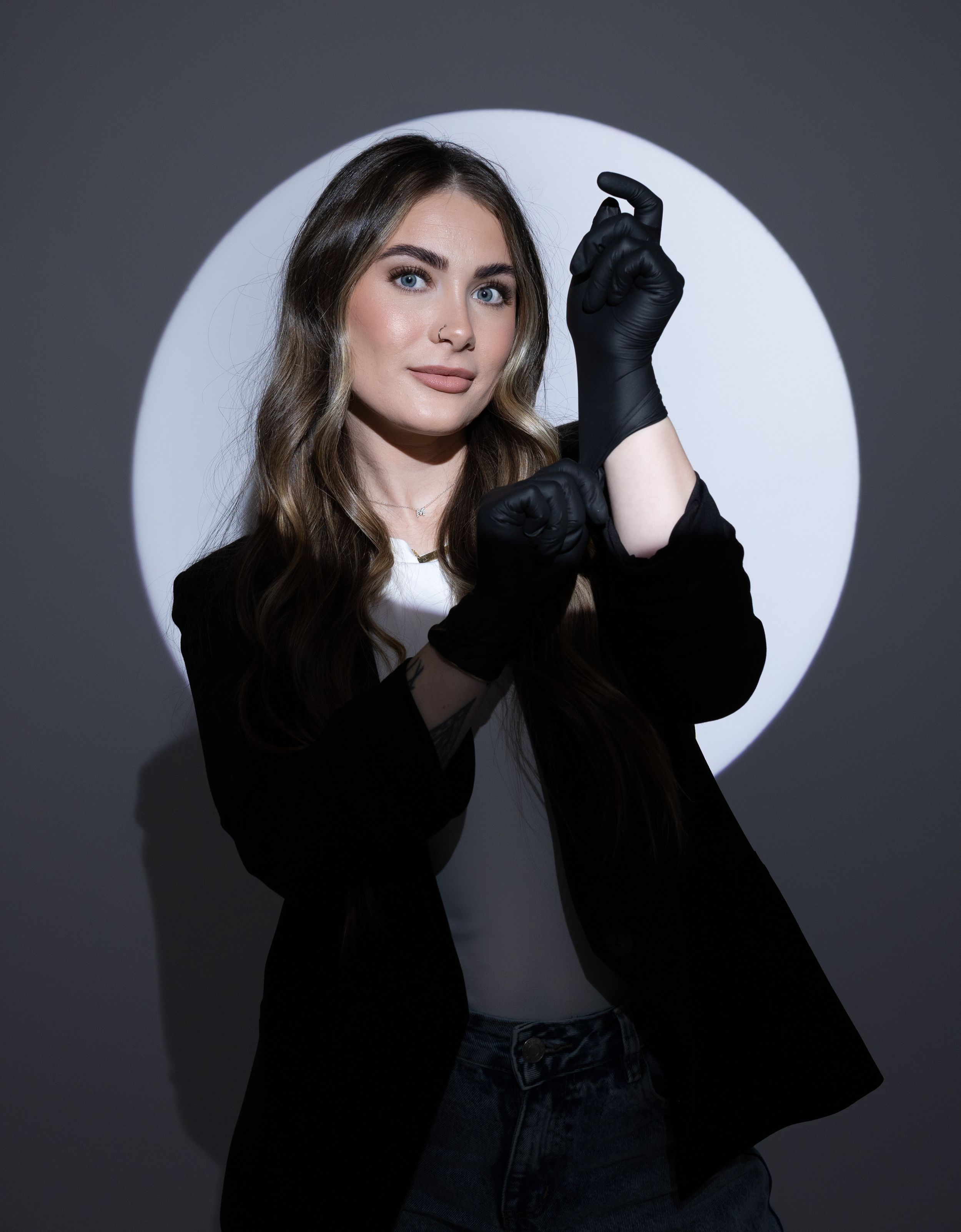 Young woman in black blazer and gloves poses confidently against circular light backdrop.