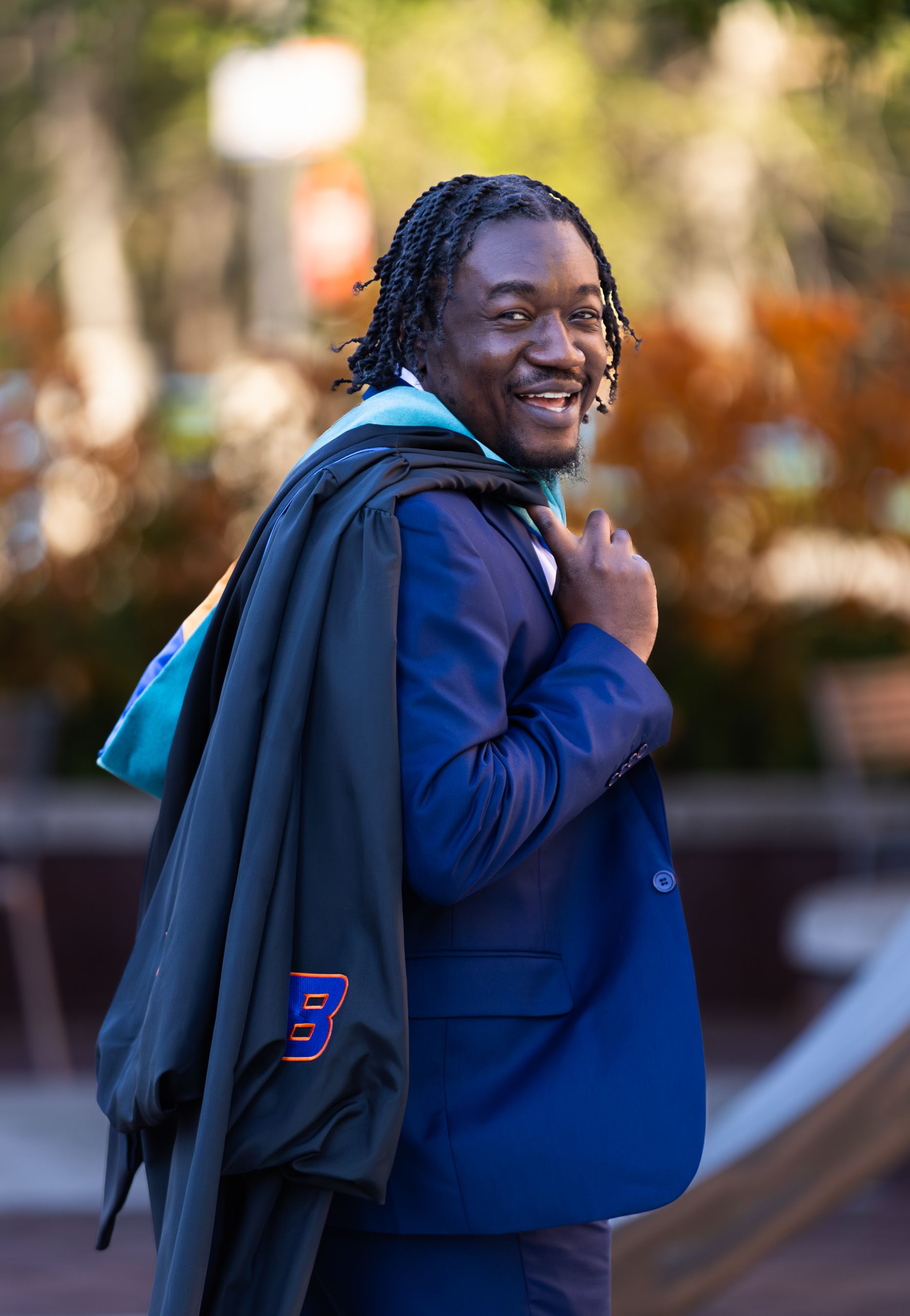 Smiling man in blue suit with graduation gown over shoulder posing outdoors on campus.