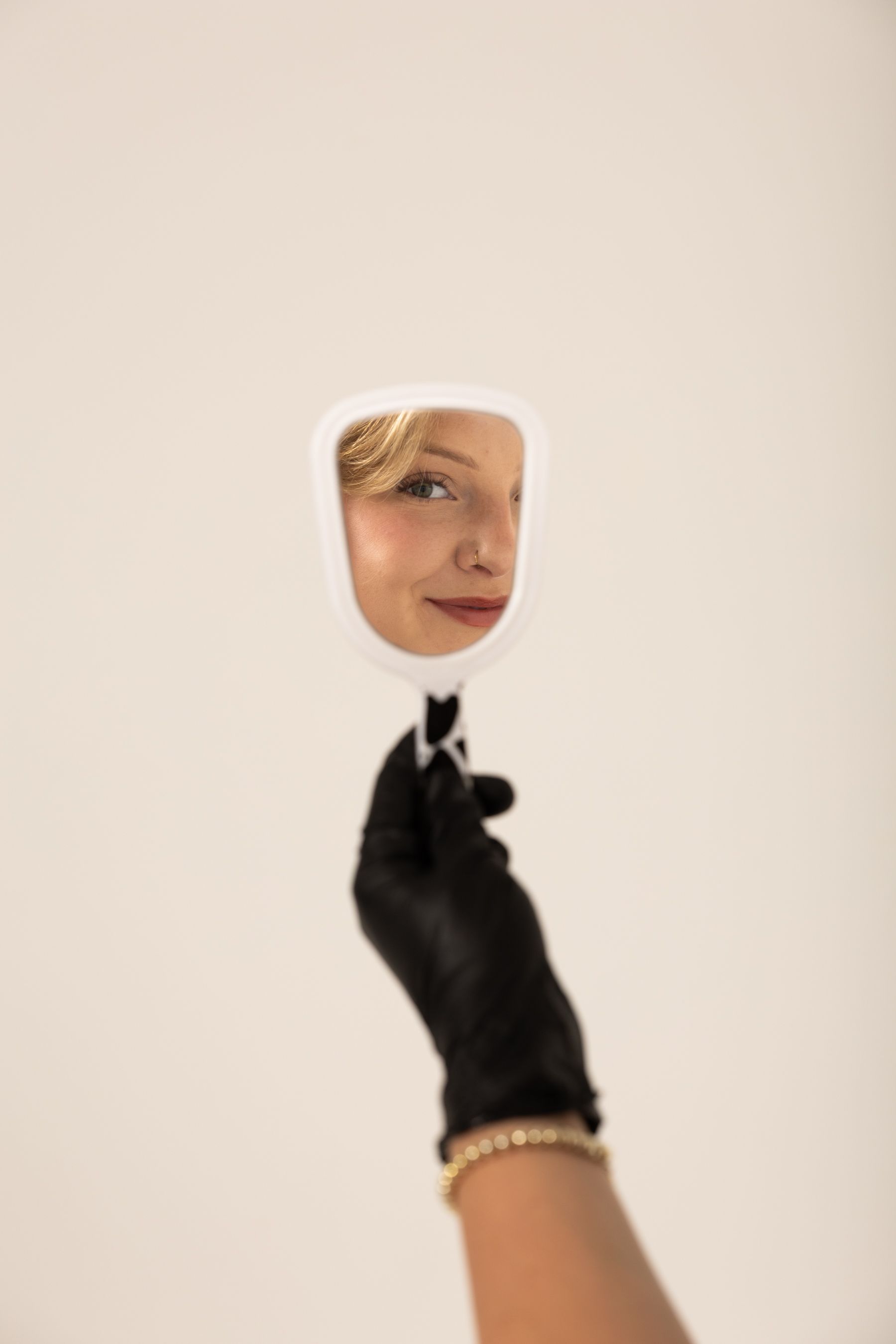 Hand in a black glove holding a small white mirror reflecting a blonde womans smiling face.