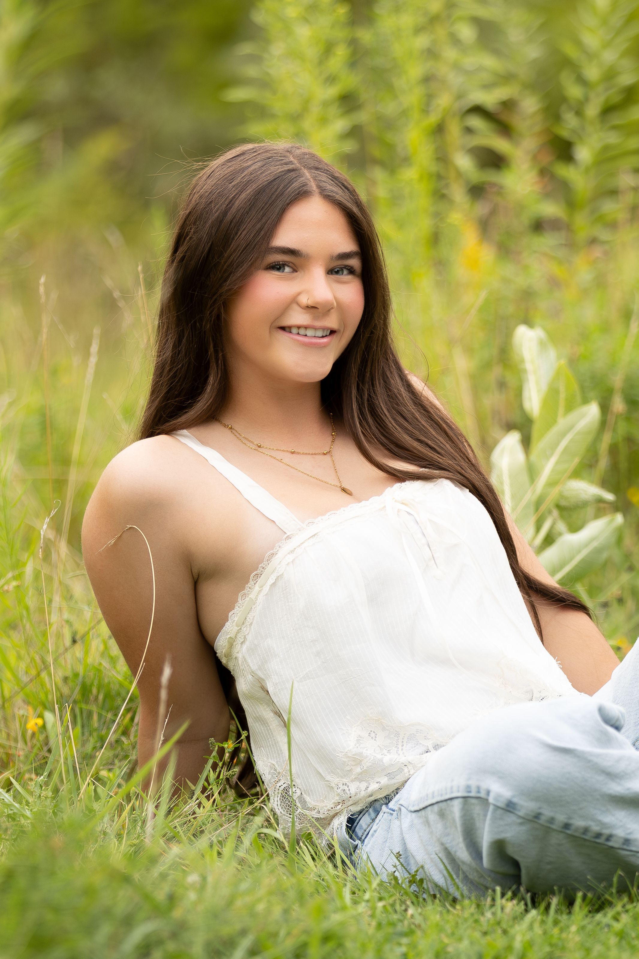 A smiling teenage girl with long brown hair sits in a lush green meadow wearing a white top and jeans.