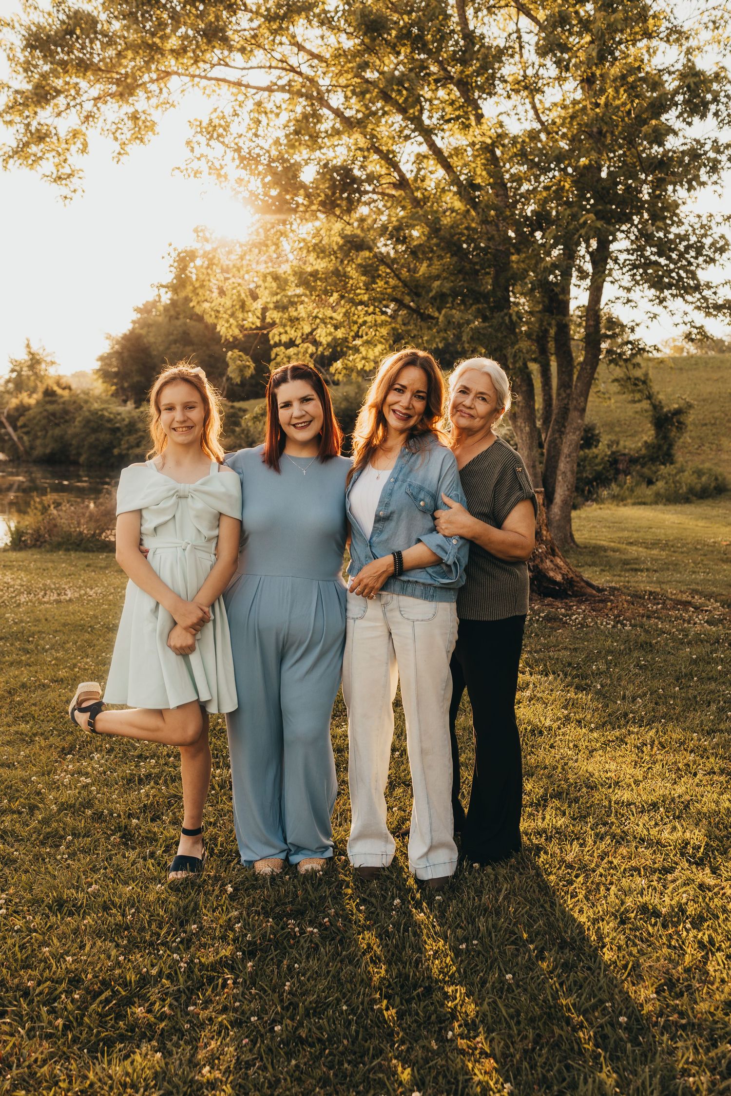 Four women smiling together outdoors at golden hour, posing on green grass with sunlit trees in the background.
