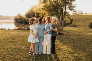 Four women smiling and embracing outdoors at golden hour near a lake with lush green grass and trees.