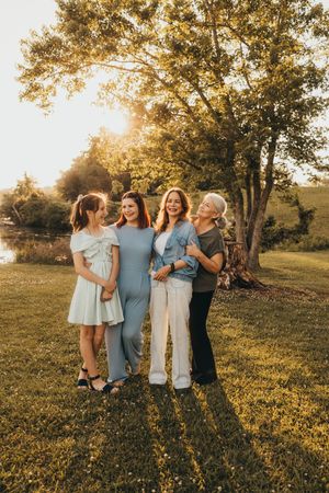 Four women of different ages smiling together outdoors in golden hour sunlight near a large tree.