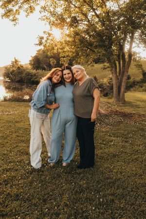 Three women smiling and embracing outdoors at golden hour near a pond surrounded by lush green trees.